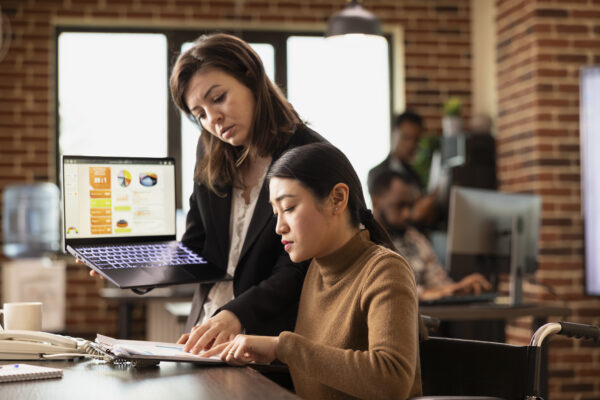 Asian employee at desk participates in business discussions while managing her recovery in modern office. Businesswoman stands with laptop, presenting marketing insights to colleague in wheelchair. Salesforce Certification Training